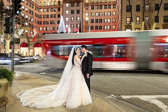 Couple portrait of bride and groom embracing with a forehead kiss, long veil and tuxedo on a city crosswalk as a streetcar blurs past at night