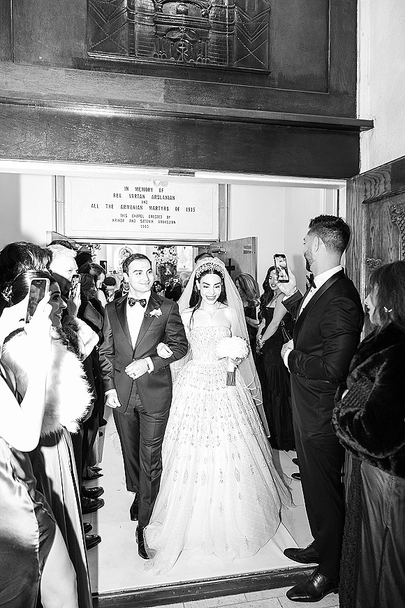Wedding recessional as bride and groom walking out church doorway, bride in tiara and veil with bouquet, guests cheering and filming on phones