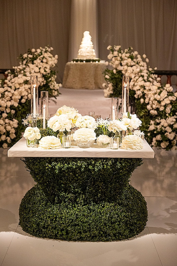 Wedding cake table set as a wedding cake display with tiered cake, white floral arrangements, and pillar candles before draped curtains