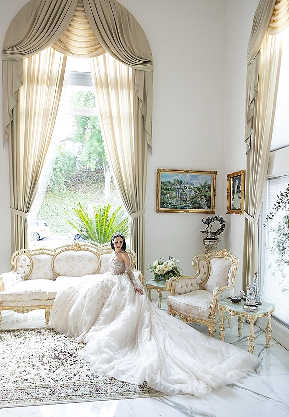 Bridal portrait of a strapless ball gown bride seated on a vintage sofa, long train flowing in a sunlit sitting room by large windows