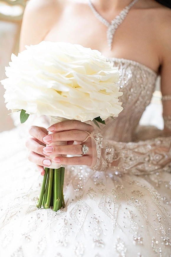 Bridal bouquet of white blooms held by the bride, bridal bouquet wrap and emerald cut engagement ring visible in soft indoor light
