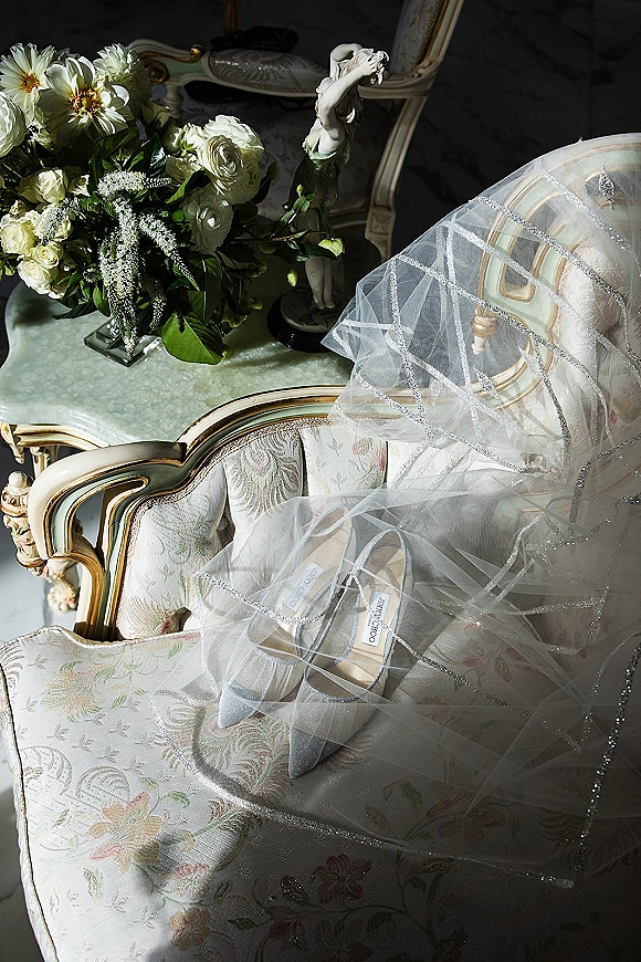 Bridal shoes in a wedding shoe flat lay, pointed toe heels draped with a sparkly tulle veil beside white roses on a vintage armchair