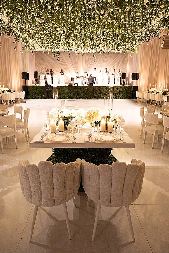 Wedding reception decor with a hanging floral ceiling, candlelit long head table, and white dance floor in a draped reception hall.