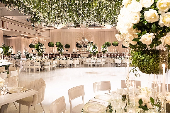 Reception ballroom decor with white floral centerpieces and taper candles on banquet tables beneath chandeliers and hanging greenery