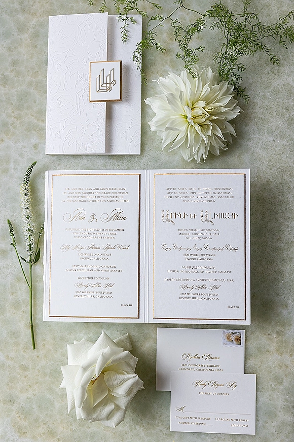 Invitation suite wedding invitation flat lay with embossed white and gold cards, monogram and RSVP, postage stamp, and white dahlia on stone surface