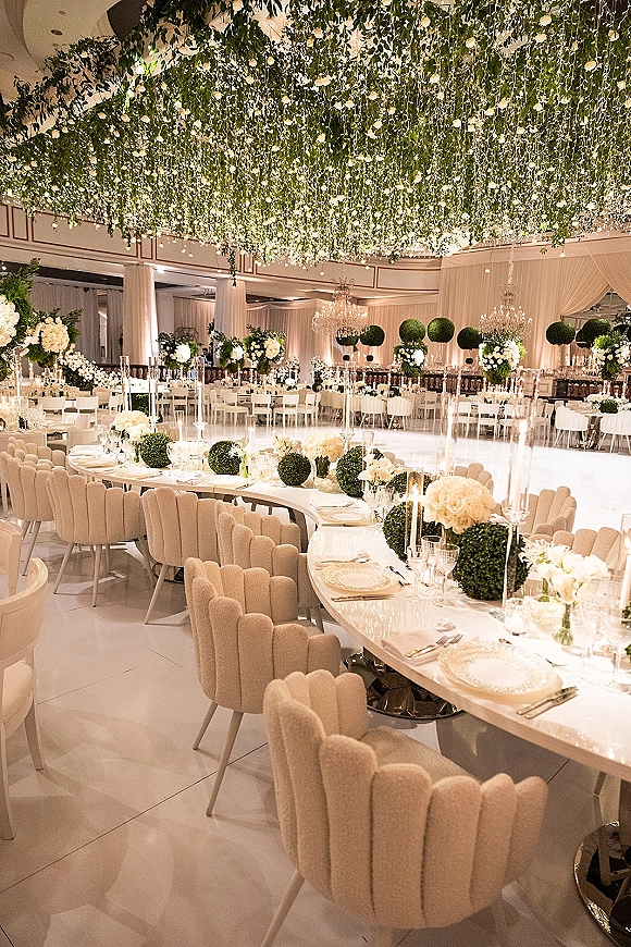 Reception decor with hanging greenery and white roses over candlelit tables, crystal chandeliers, and a white dance floor in a ballroom