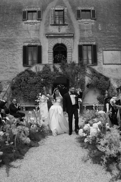 Wedding recessional as bride and groom walk up aisle with bridal veil and bouquet, guests applauding in ivy-covered stone villa courtyard