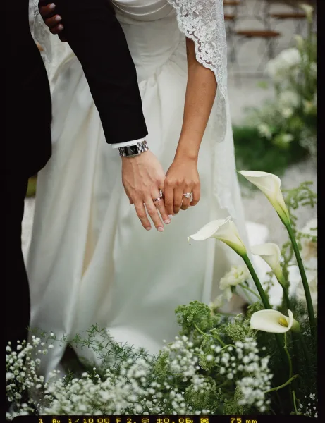 Wedding hands clasped with wedding rings close up, bride’s lace sleeve and groom’s tuxedo cuff and watch by calla lilies on stone steps