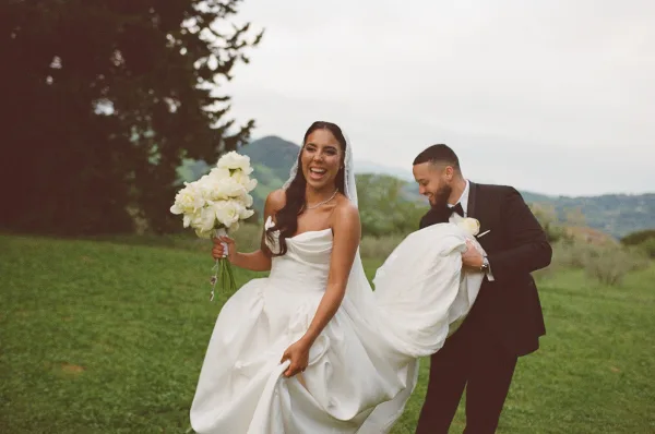 Couple portrait outdoors, bride and groom laughing as he holds her dress and she carries a bouquet in a mountain meadow under clouds