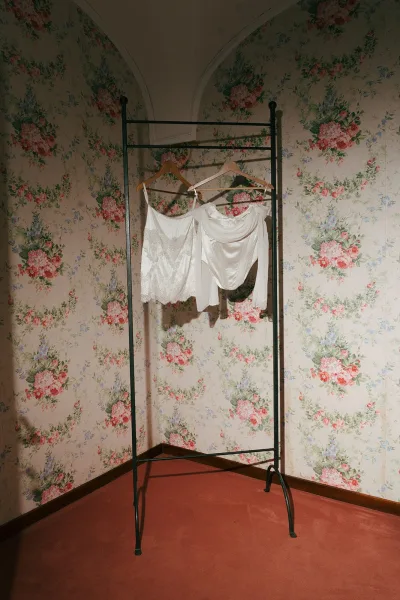 Wedding dress detail with lace and satin hangs on wooden hangers from a garment rack against floral wallpaper and an arched niche