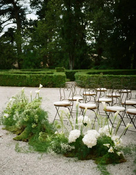 Ceremony aisle decor with white flowers, baby's breath, and greenery on moss beside wrought iron chairs in a gravel garden courtyard