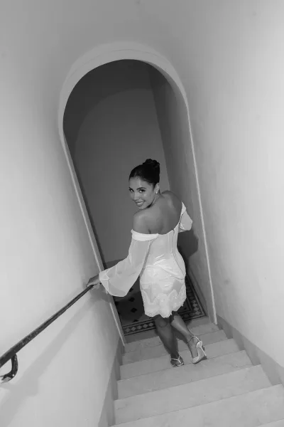 Bridal portrait of a bride in a short wedding dress looking back on a staircase, hand on rail, with an arched doorway behind her