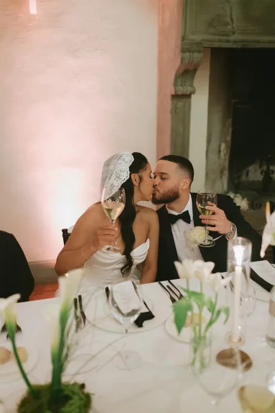Wedding kiss moment as bride in lace veil and groom in tuxedo lean over sweetheart table, holding champagne amid candlelight and white florals by fireplace