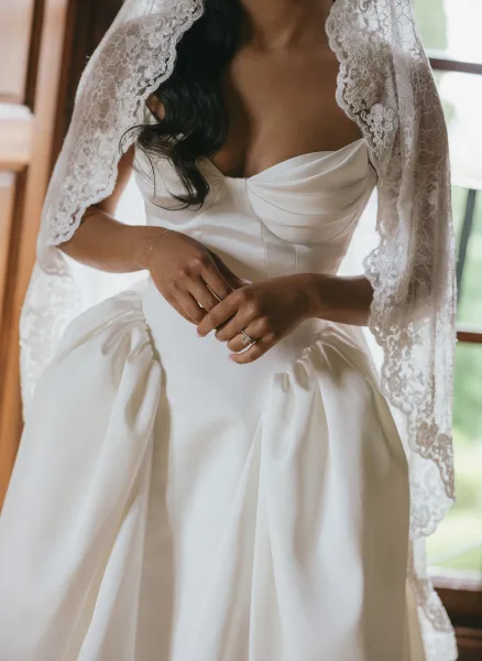 Wedding dress close-up of a strapless satin wedding dress with lace veil, engagement ring and bracelet, lit by a window with greenery outside