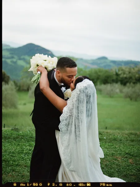 Wedding kiss as bride and groom embrace, her lace veil trailing and white bouquet in hand against misty mountains and green hillside