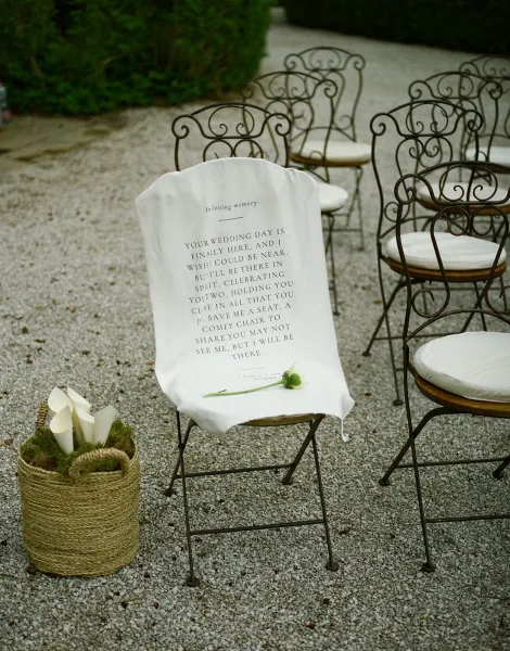 Memorial chair sign draped over a fabric cover with a printed message and small flower bud on a wrought iron ceremony chair on gravel amid greenery