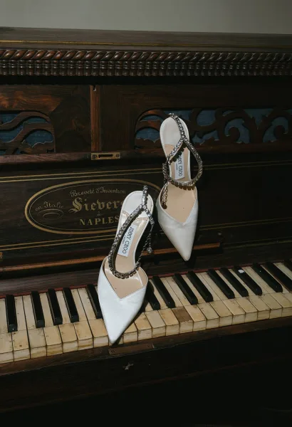 Wedding shoes, bridal heels in white satin with rhinestone ankle straps, resting on vintage piano keys against dark wood backdrop