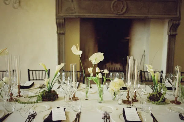 Reception tablescape with wedding table setting, white calla lilies and anthurium in glass vases, taper candles on brass by a fireplace mantel