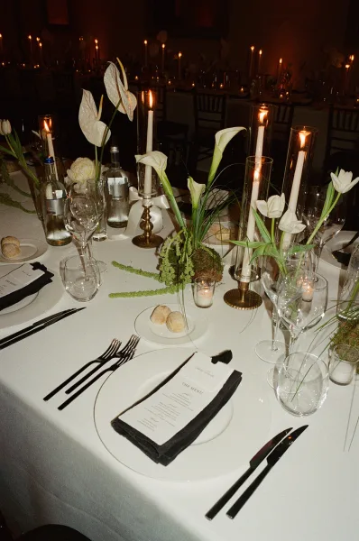 Reception tablescape with a candlelit wedding reception table, white cloth and black napkins, brass tapers and florals in a dim room