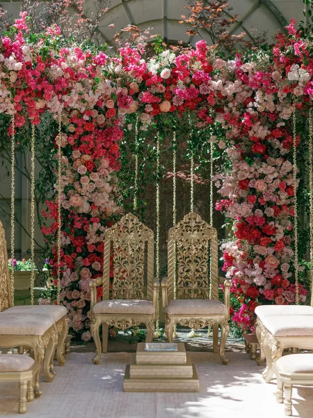 Ceremony setup beneath a floral wedding arch with gold chairs amid garden greenery, backed by a stone wall and arched trellis.