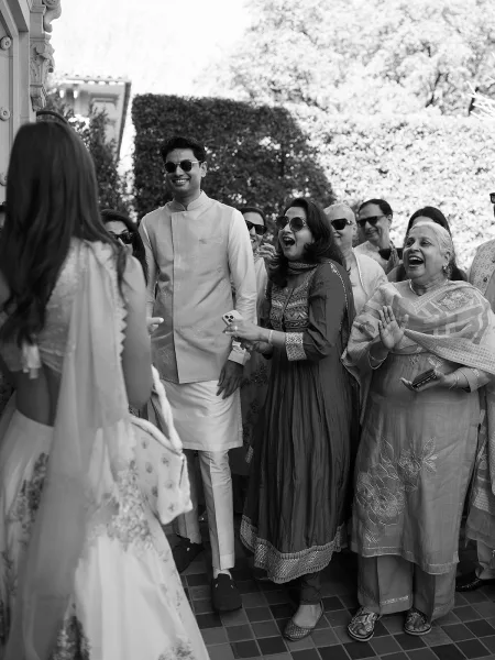 Wedding guest reaction during the bride entrance moment, with cheers and smiles, as she appears in a bridal lehenga by a garden hedge and stucco wall.