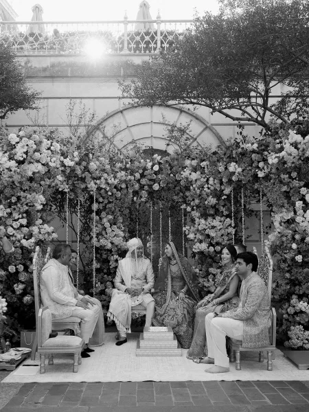 Ceremony moment at a Hindu wedding ceremony as the bride and groom sit beneath hanging flower strings, with garden trees and a stone wall behind.