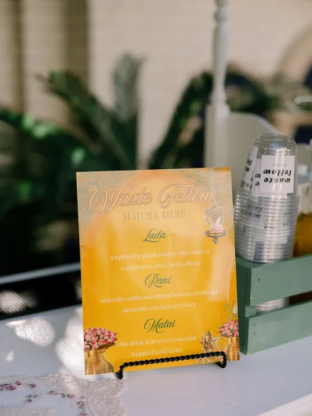 Wedding bar sign presenting a drink menu sign on a lace table runner, set against palm plants and a rustic brick wall.
