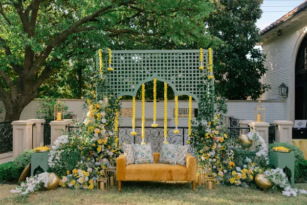 Wedding lounge decor with outdoor lounge seating centered on a velvet sofa, set on a grass lawn beneath garden trees and a brick wall.