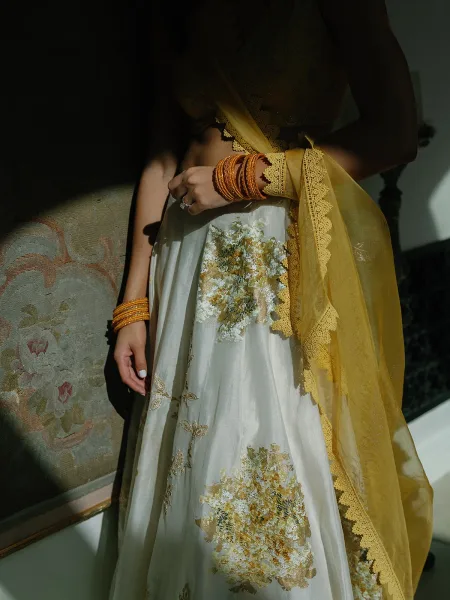 Bridal lehenga detail of a bride in a yellow dupatta, showcasing an ivory skirt’s delicate textures against a patterned tapestry wall.