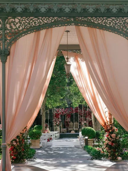 Ceremony setup with refined garden ceremony decor, a flower wall framing a raised platform beneath a pavilion amid garden trees.