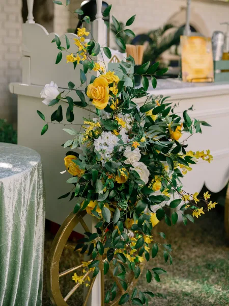 Wedding bar decor for cocktail hour bar styling with yellow roses adorning the setup on a grass lawn beside a brick wall and palm leaves.