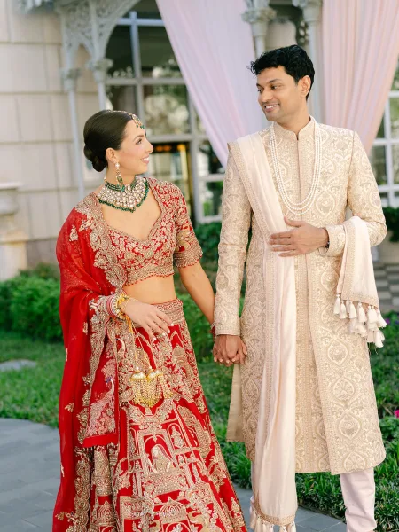 Couple portrait of bride and groom holding hands, the bride in a red bridal lehenga, set against garden greenery and a light building facade.