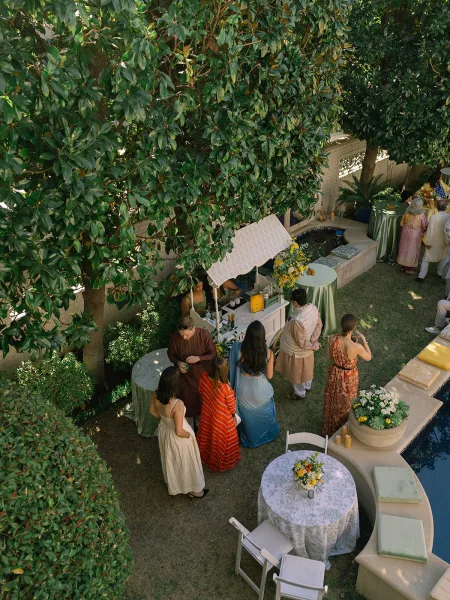 Cocktail hour setup for an outdoor cocktail reception with a bar cart by the swimming pool on a grass lawn framed by garden trees and hedges.
