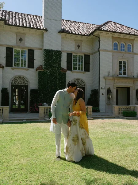 Wedding kiss portrait of an Indian wedding couple on an estate lawn, her sheer yellow dupatta floating as ivy-clad walls and blue sky frame them.