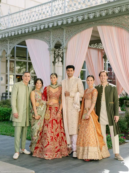 Wedding party portrait of an Indian wedding group on a wrought-iron veranda, bride in a bridal lehenga with family in coordinated attire.