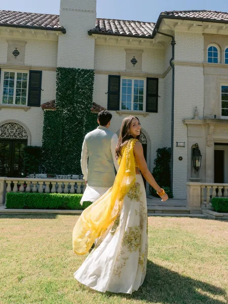 Couple portrait featuring a bride in lehenga and her partner smiling on the lawn before an ivy-clad mansion facade under a clear blue sky.