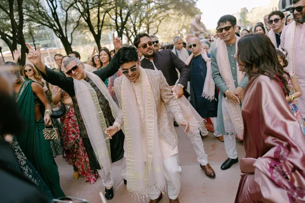 Baraat procession flows into a lively wedding street dance, sherwani-clad guests celebrating along a tree-lined pavement beside city buildings.
