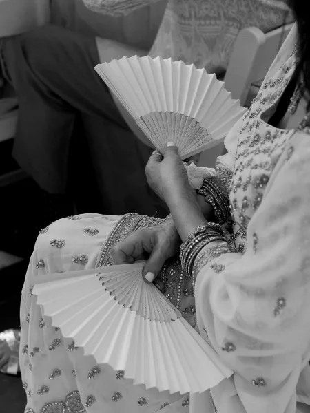 Wedding ceremony fans double as hand fan wedding favors, held beside an embellished sari amid guest seating and patterned textiles.