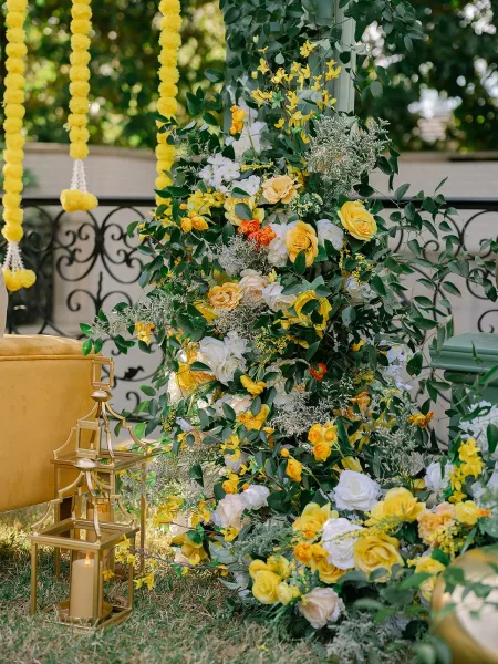 Ceremony flowers in yellow and white florals create a lush display, accented with marigold garlands against garden trees and wrought iron railing.