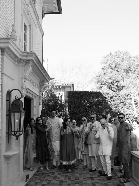 Wedding group portrait of wedding guests cheering, some in kurtas, on a tiled patio by a building facade and garden hedges.