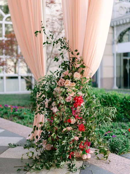 Wedding arch flowers frame a draped ceremony arch with roses, set in a courtyard with garden beds and a stone walkway.