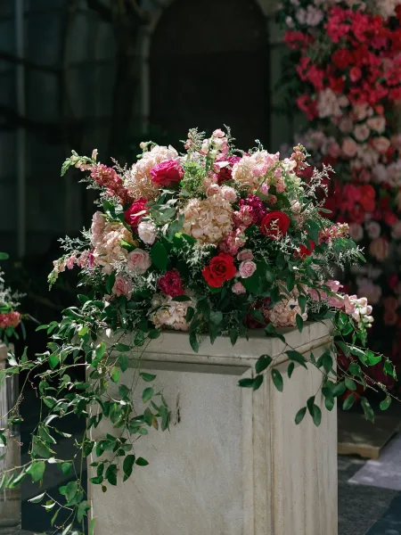 Ceremony flowers styled as a pedestal floral arrangement with lush roses under a stone archway and garden greenery backdrop.
