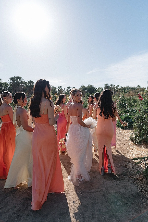 Bridesmaid group photo of bride with bridesmaids walking on a sunny garden path, holding bouquets in pink and coral dresses under palms