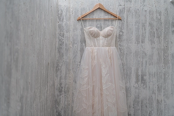Wedding dress on a wooden hanger, strapless wedding dress with floral lace applique and tulle skirt against a textured concrete wall