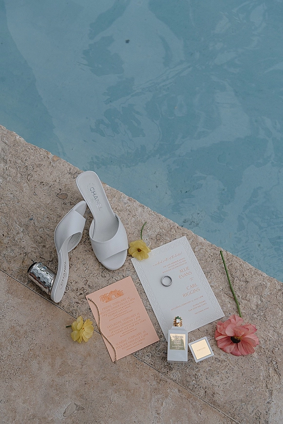 Wedding flat lay with wedding invitation flat lay, white heeled sandals, rings, perfume, and flowers on a stone pool deck by blue water