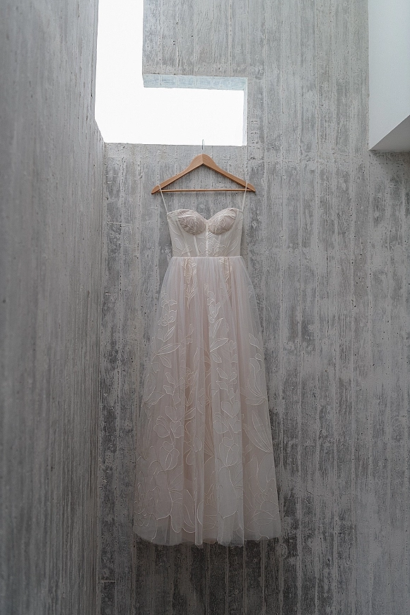 Wedding dress hanging on a wooden hanger, strapless wedding dress with floral embroidery and tulle skirt against a concrete wall and skylight window