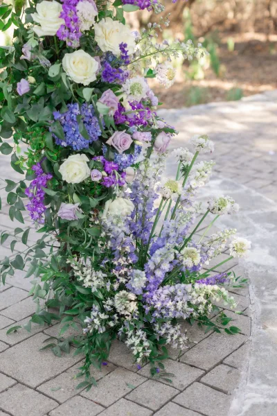 Wedding ceremony florals with ceremony aisle flowers in white and lavender roses, blue hydrangea, and greenery beside a stone walkway outdoors