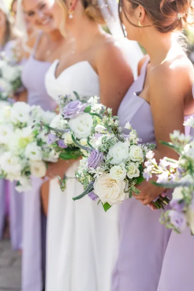 Bridesmaid bouquets with lavender and white bouquet of white roses, lavender flowers, and greenery, held in a sunlit outdoor walkway with guests blurred behind