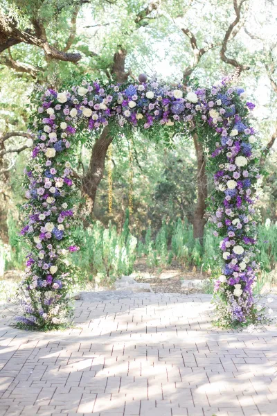 Wedding ceremony arch with purple and white roses and hydrangeas, framed by greenery garland in an outdoor garden with trees and brick walkway