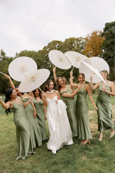 Bride with bridesmaids in sage green dresses holding white paper parasols, posing in a grassy field with trees under overcast sky